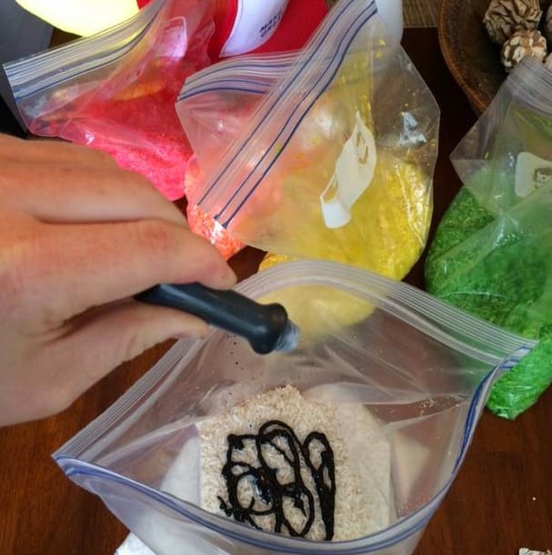 a hand squeezes a bottle of blue food coloring into a Ziploc bag of white rice.