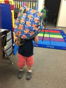 A little boy stands in the classroom with a colorful backpack on his head.