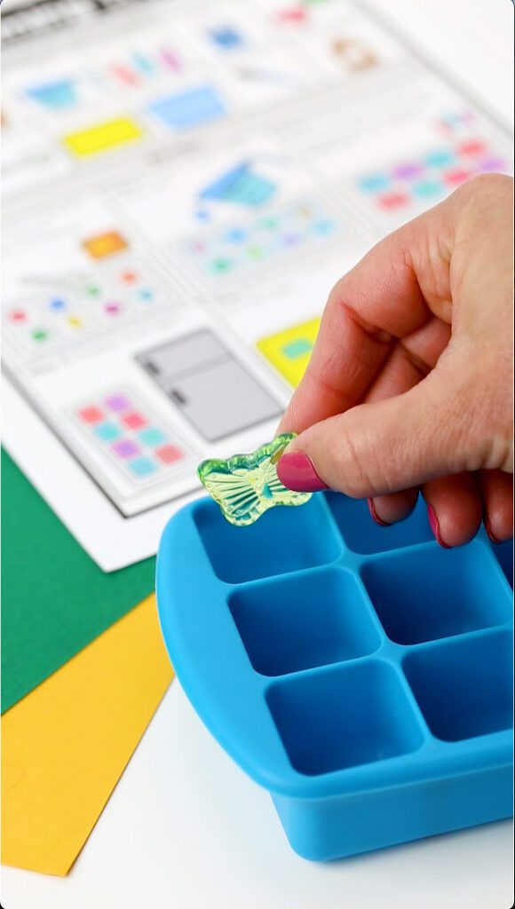 woman adding a butterfly charm to a blue plastic ice cube tray for a buried treasure visual recipe instructions for a science experiment for preschool and special education students. 