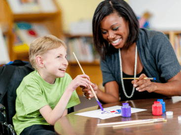 A teacher helps a child in a wheelchair to paint a picture