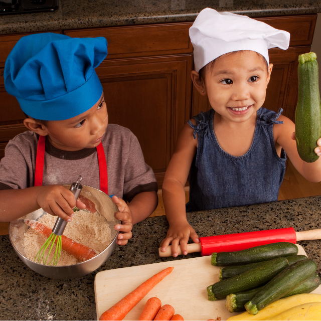 Two children in the kitchen, wearing chefs hats. The little girl is holding up a courgette and the little boy is trying to stir a whole carrot into a bowl of flower with a whisk.

