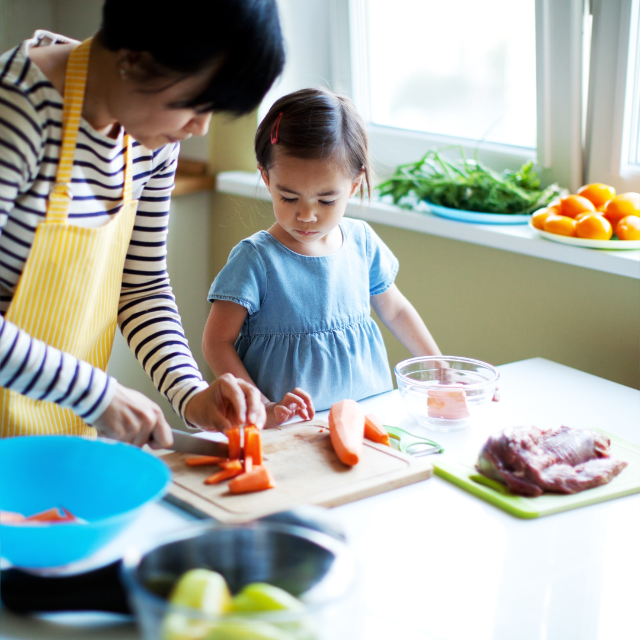 A girl stands at the kitchen counter. She is watching a woman cutting a carrot on a chopping board. In front of them are bowls with other ingredients but they are blurrred and unclear.
