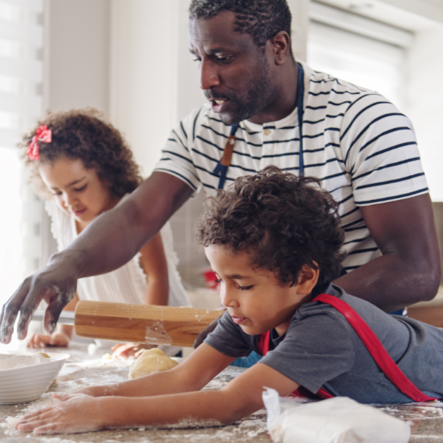 A male adult and two children are cooking together at the kitchen counter. The man is reaching for some flour to sprinkle on the dough he is about to rool out. The boy is playing in the flour, and the girl appears to be cutting shapes out of dough.
