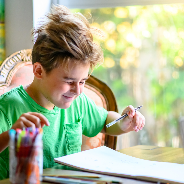 A smiling homeschooled child, holding a pen, working independently at the table.