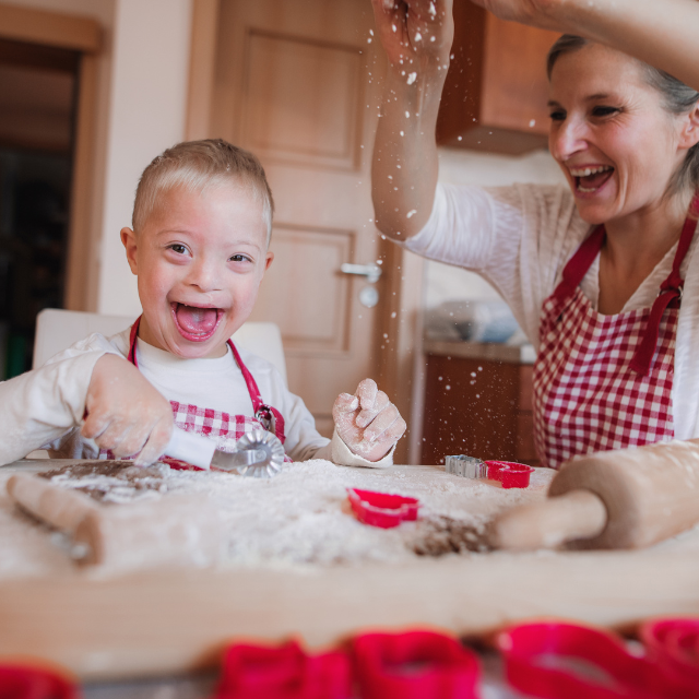 Child and mother baking in the kitchen. The mother is sprinkling flour from a haight, onto the counter. Both are laughing.