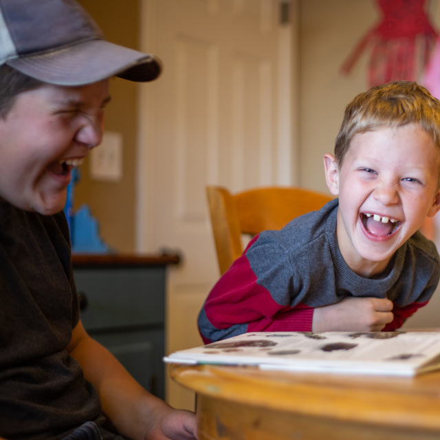 Two children at a table. They are looking at a book together and laughing.
