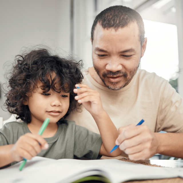 Homeschooling father and child with pens writing in a workbook at the table.