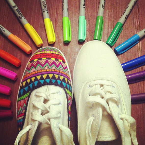 Two white tennis shoes, one undecorated and the other decorated with vibrant, multi-color geometric patterns. The shoes have a semi-circle of sharpies aranged around them.