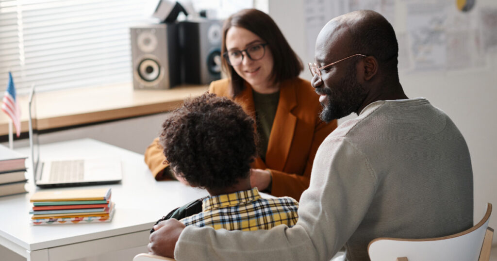 a smiling Teacher meeting with a happy African American parent and student at a desk in the classroom.