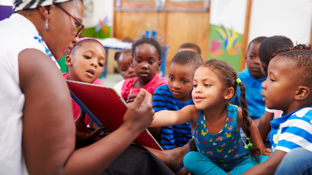 African American teacher in a white short sleeved blouse sitting on the floor reading to her early education students.