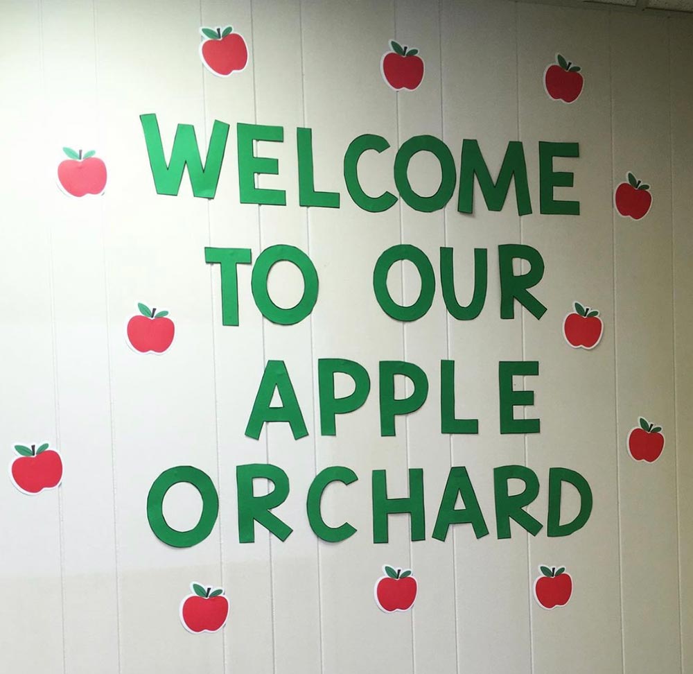 An elementary school classroom with a "Welcome to Our Apple Orchard" bulletin board.