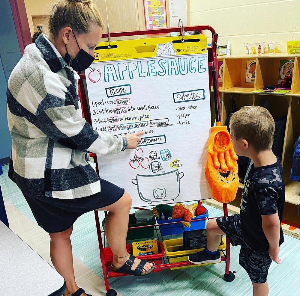 Student participates in a slow-cooker applesauce activity by pouring an ingredient into a black slow cooker with adult physical support.