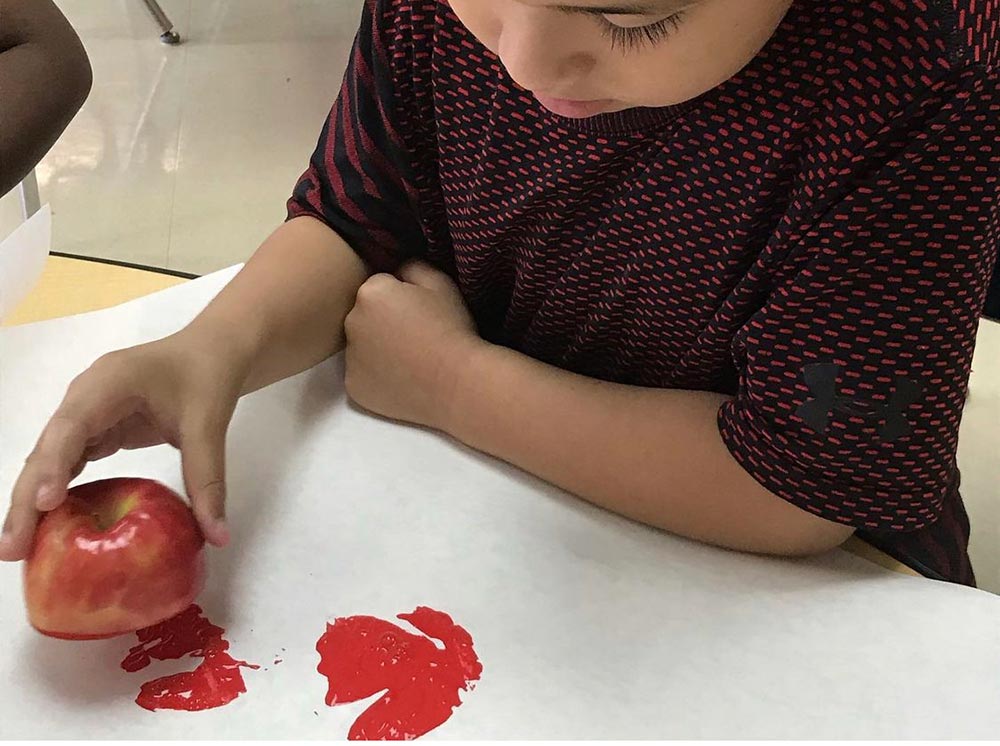 Student uses a red apple half dipped in yellow paint to stamp prints onto white paper during a classroom apple art activity.