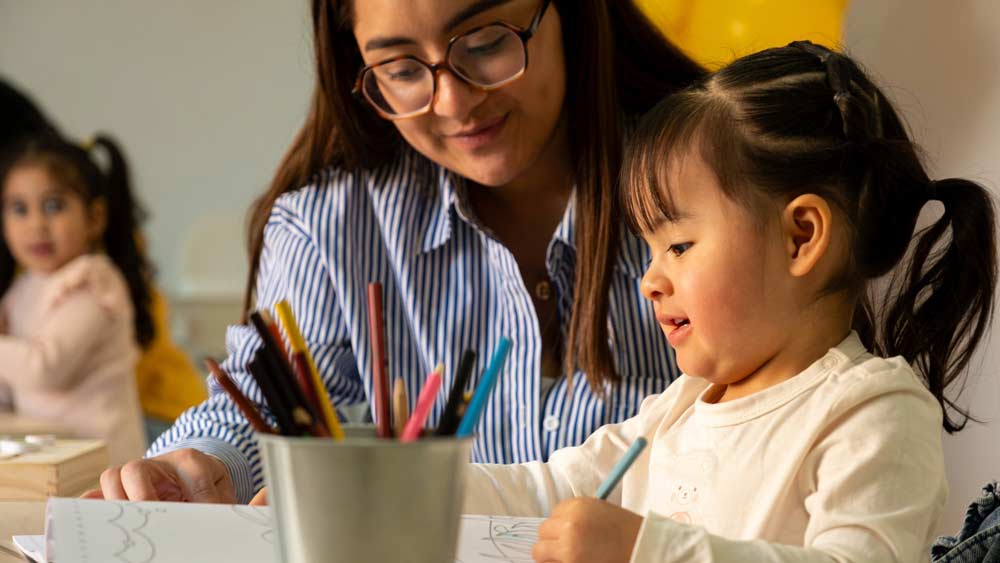 A teacher sits beside a young student as she colors a worksheet with a blue pencil at a classroom table.